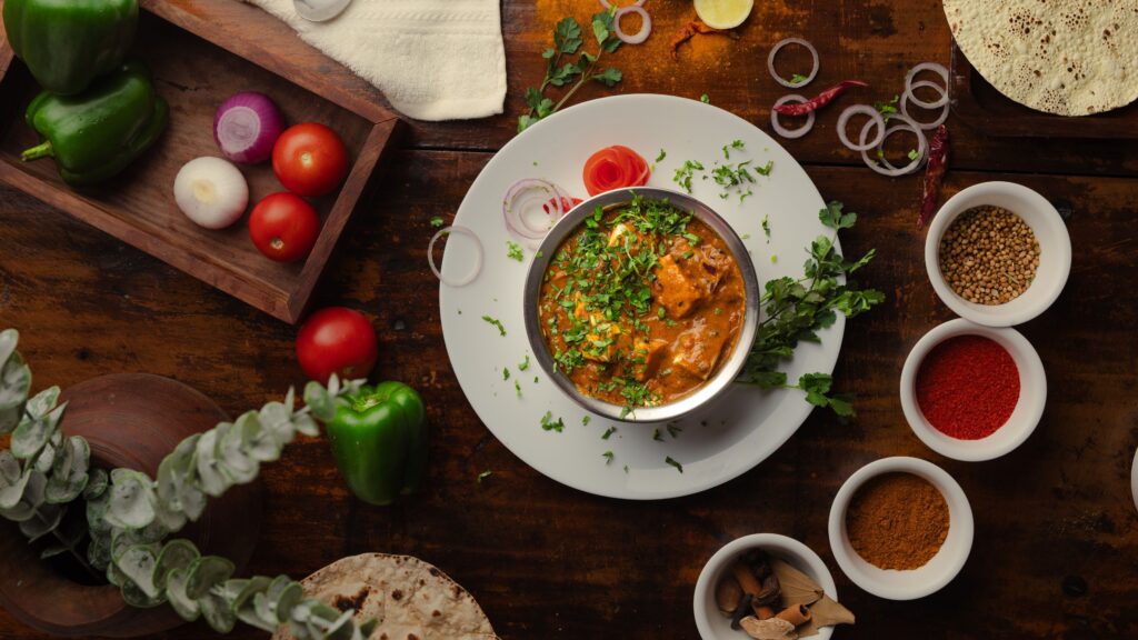 A vibrant Indian meal surrounded by spices and fresh vegetables, shot from above.